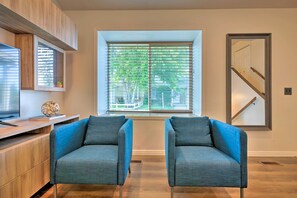 Ein modernes Wohnzimmer mit einem großen Fenster, zwei blauen Sesseln, einem Holzschrank und einem Flachbildfernseher.