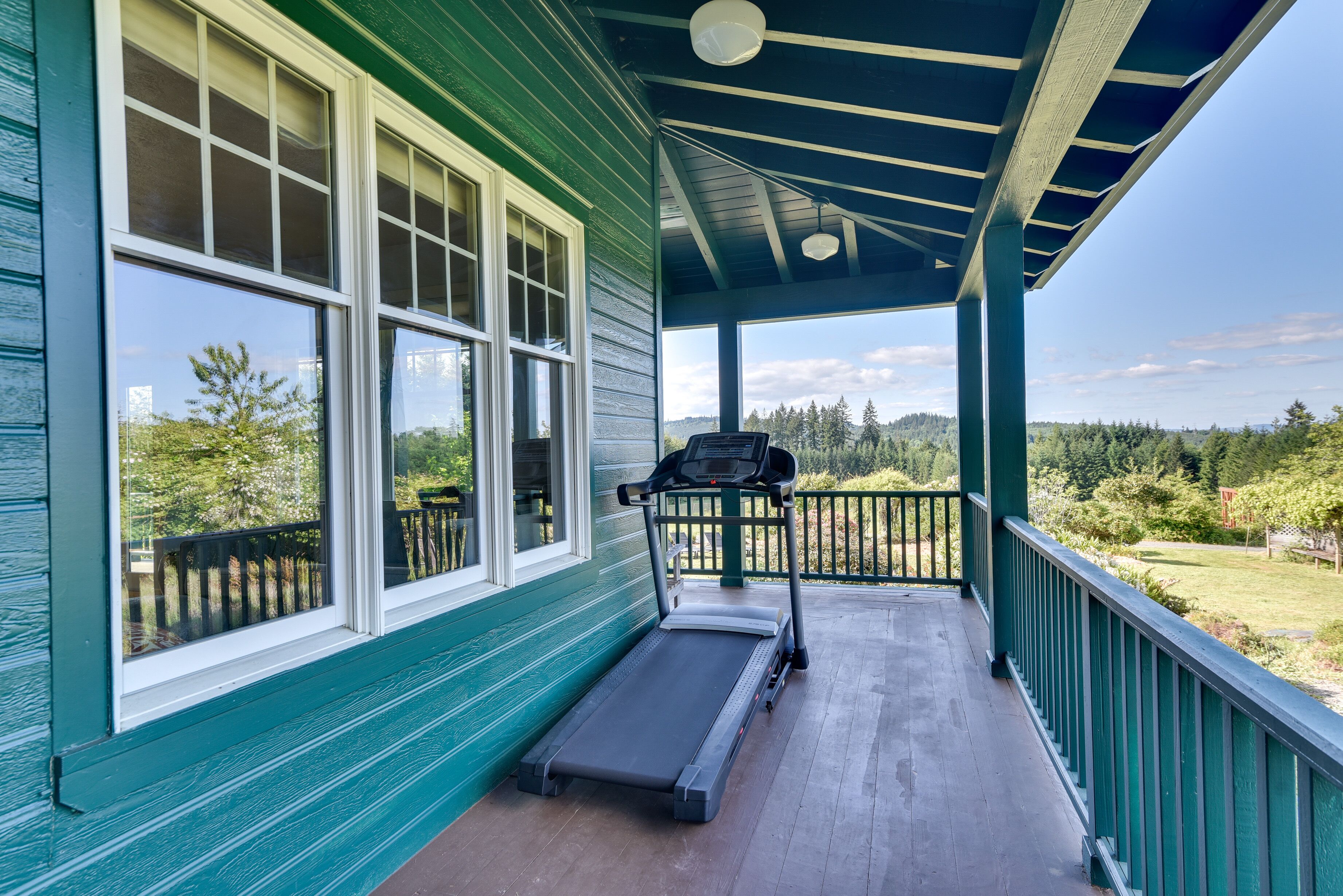 A covered porch with a treadmill, large windows, and a view of a green landscape.