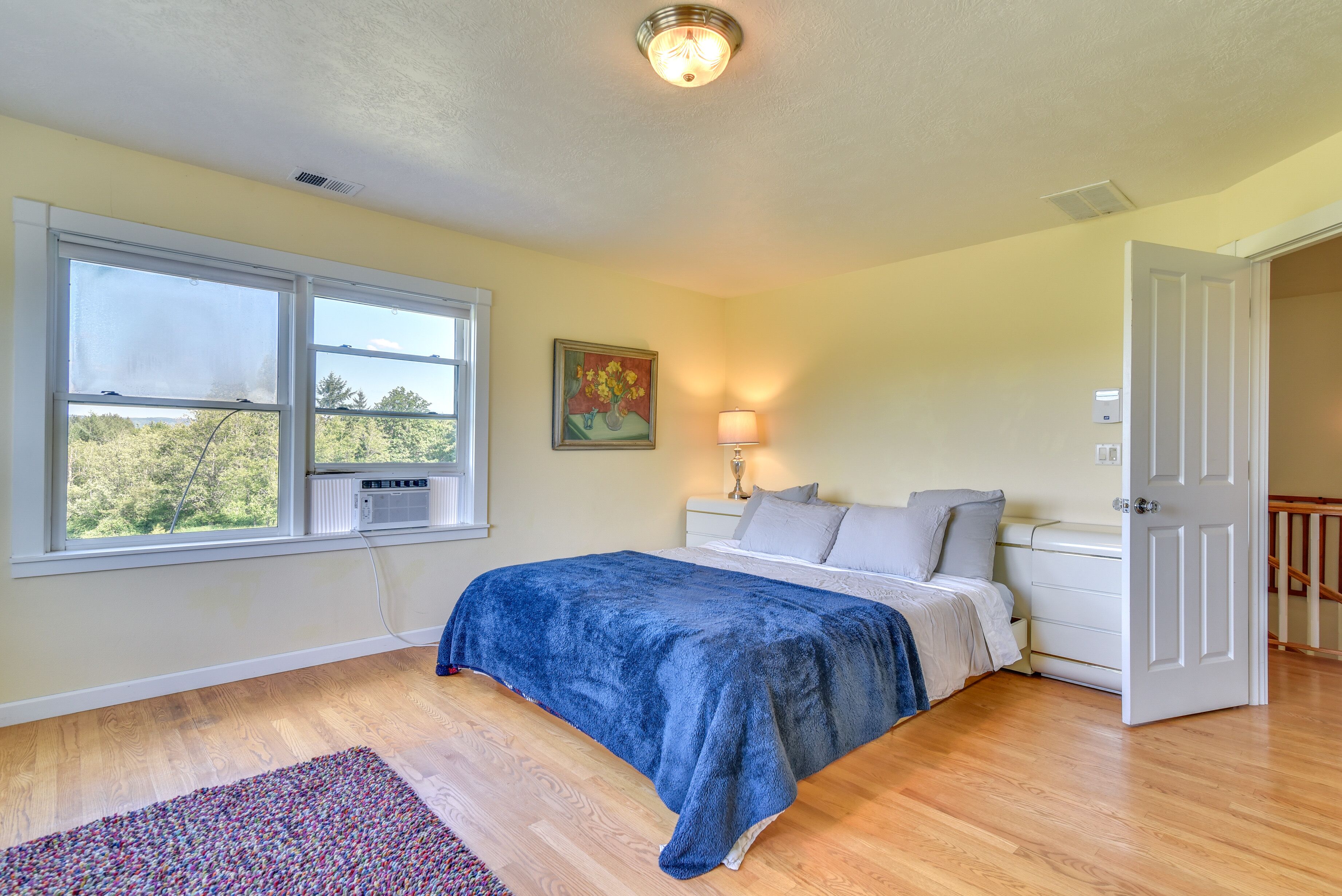 A bedroom with a large window, a bed with a blue blanket, a bedside lamp, and a door to another room.