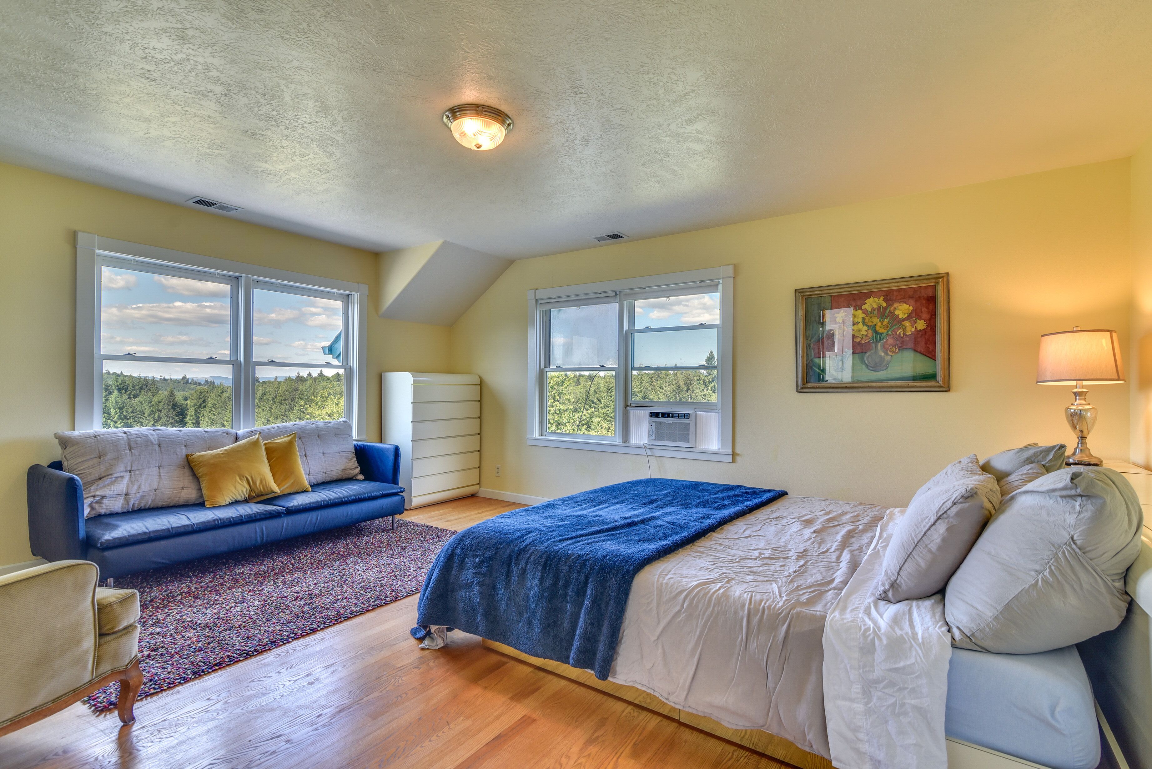 A bedroom with a bed, a blue sofa, a window with a view, and a painting on the wall.