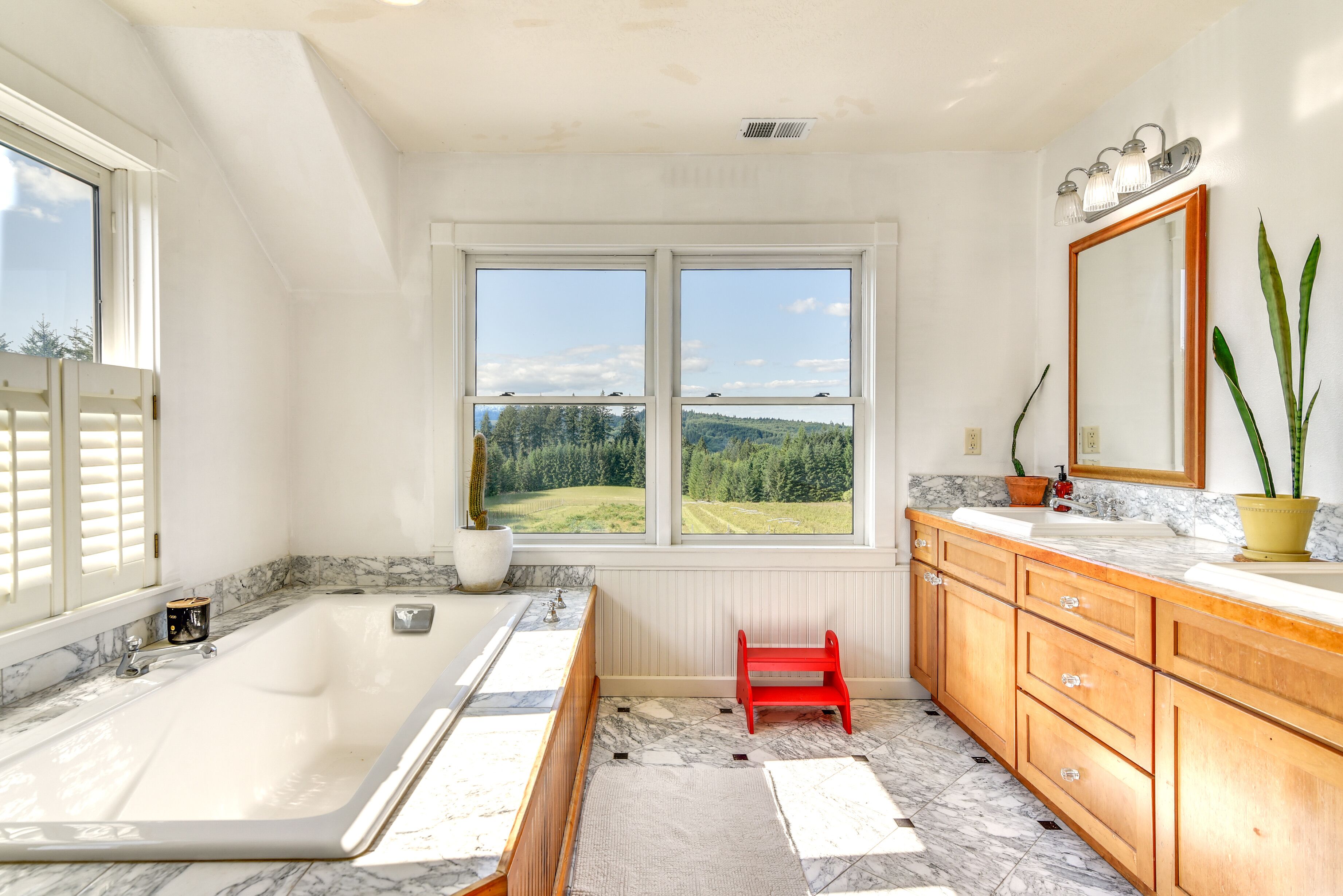 A bathroom with a bathtub, a red bench, a large window with a view of greenery, and wooden cabinets.