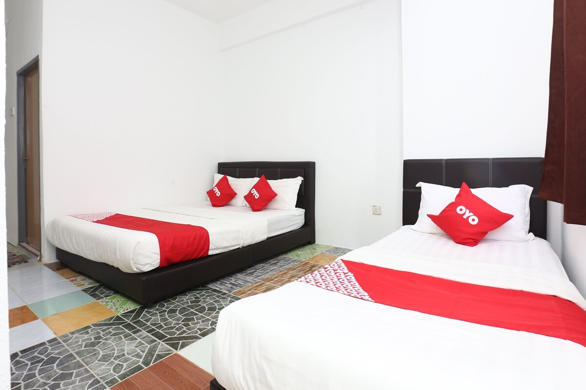 A hotel room with two beds, each with a black headboard and white bedding. There are red pillows with the logo "OYO" on them. The room has a tiled floor with a mix of patterns and colors.