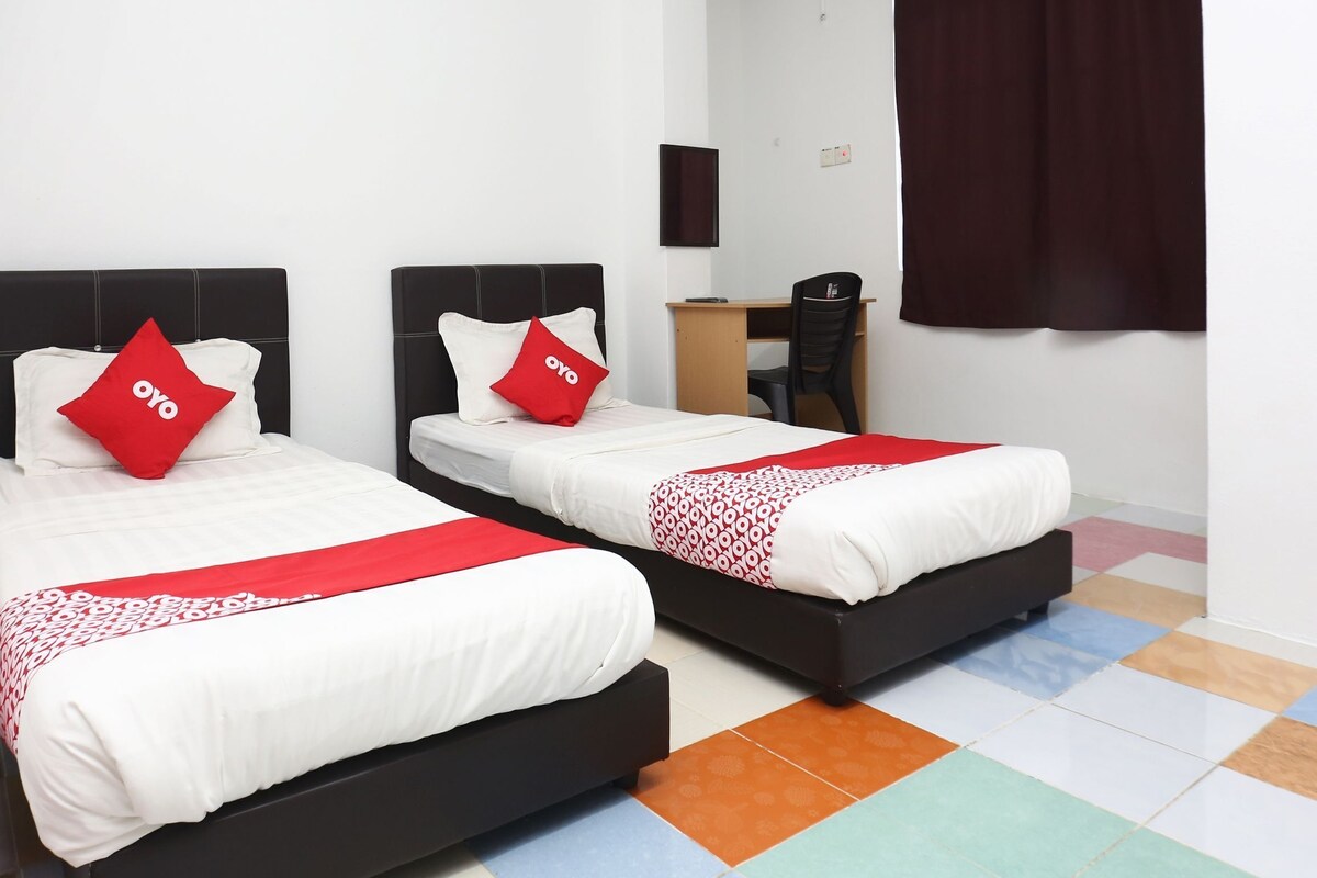 A hotel room with two beds, each with a black headboard, white bedding, and red pillows. There is a desk with a chair, a window with dark curtains, and a colorful tiled floor.