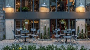 An outdoor seating area with metal chairs and tables, potted plants, and a view of the interior through large windows. - Bridges Danang Boutique Hotel (Da Nang)