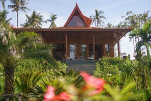 A traditional wooden building with a sloped roof, surrounded by lush greenery and palm trees. - Villa Ronggo Mayang Bali (Selemadeg)