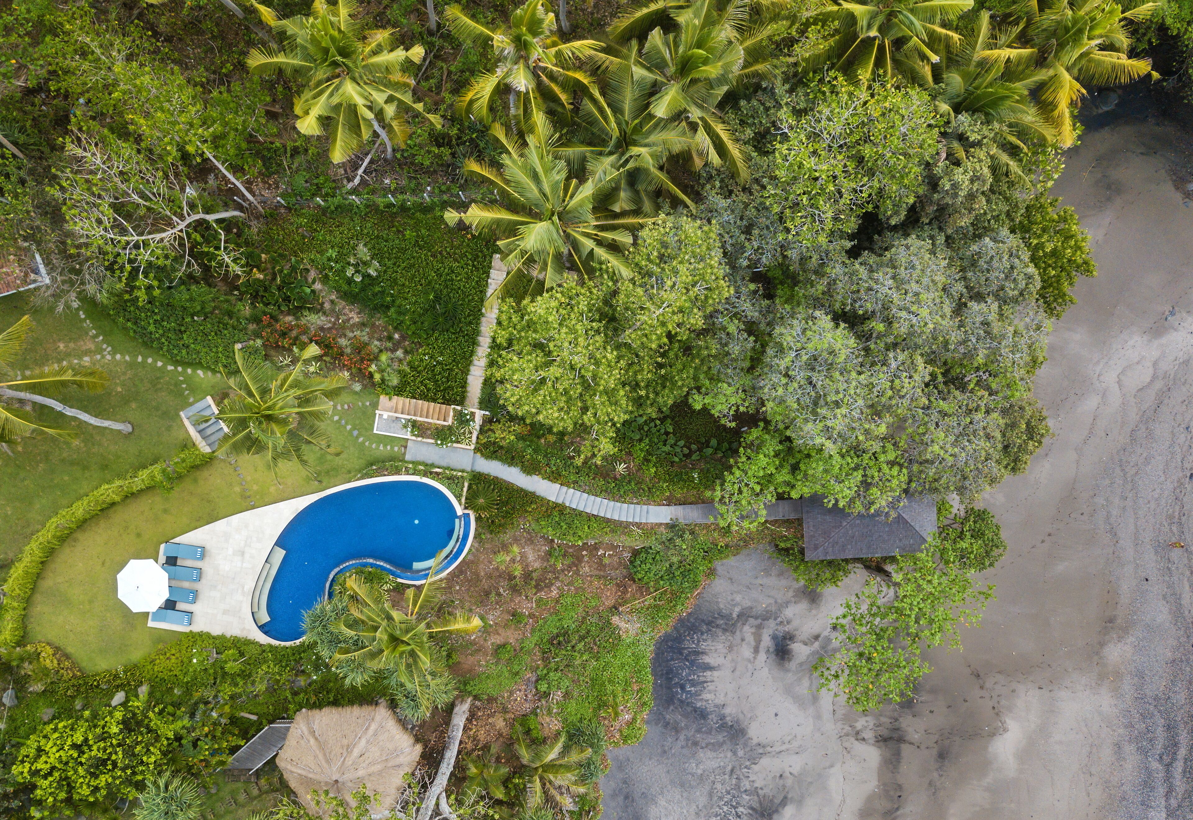 An aerial view of a resort with a swimming pool, lush greenery, and a beach.