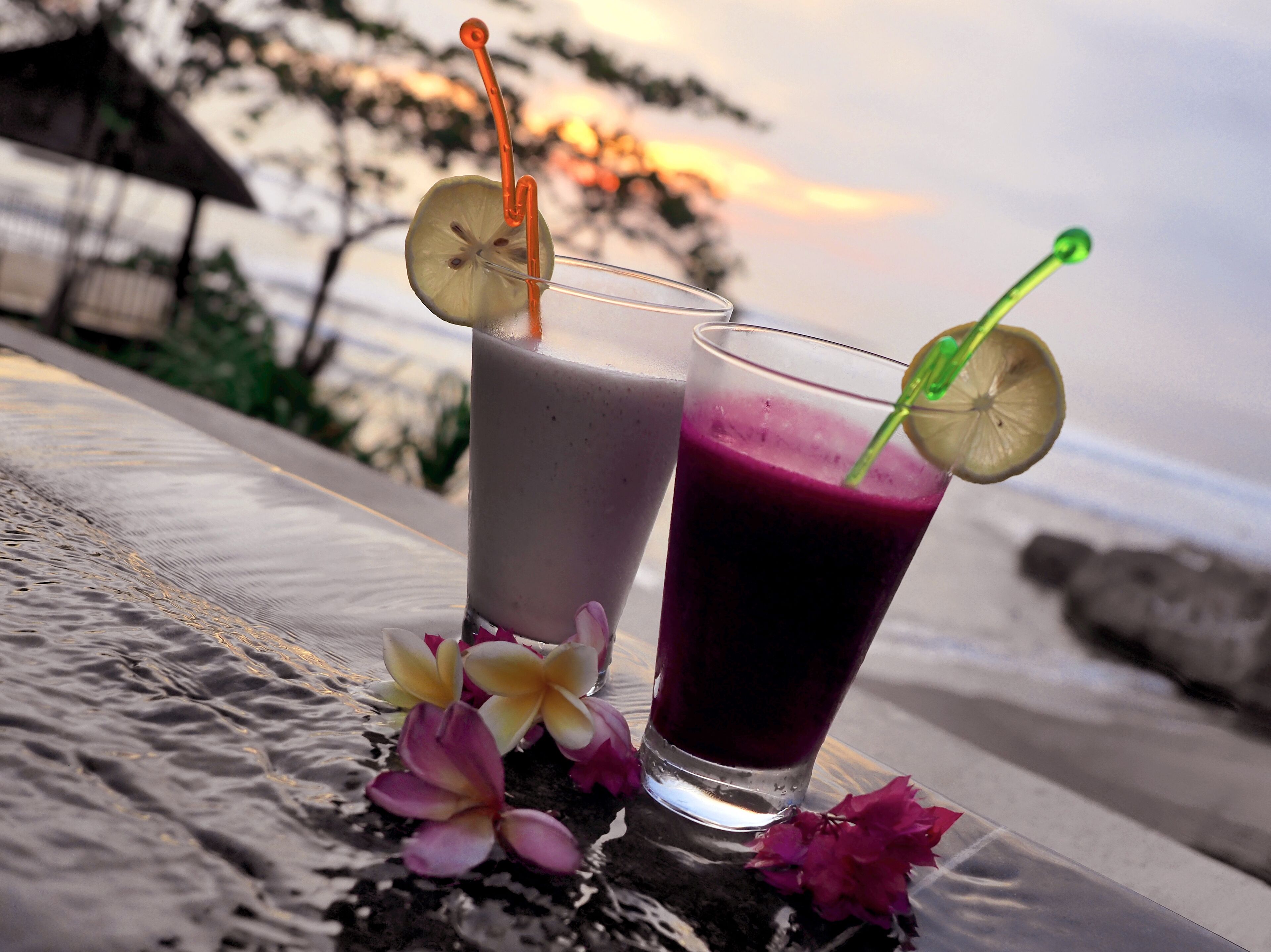 Two glasses of beverages with straws and lemon slices, placed on a table with flowers and a beach view in the background.