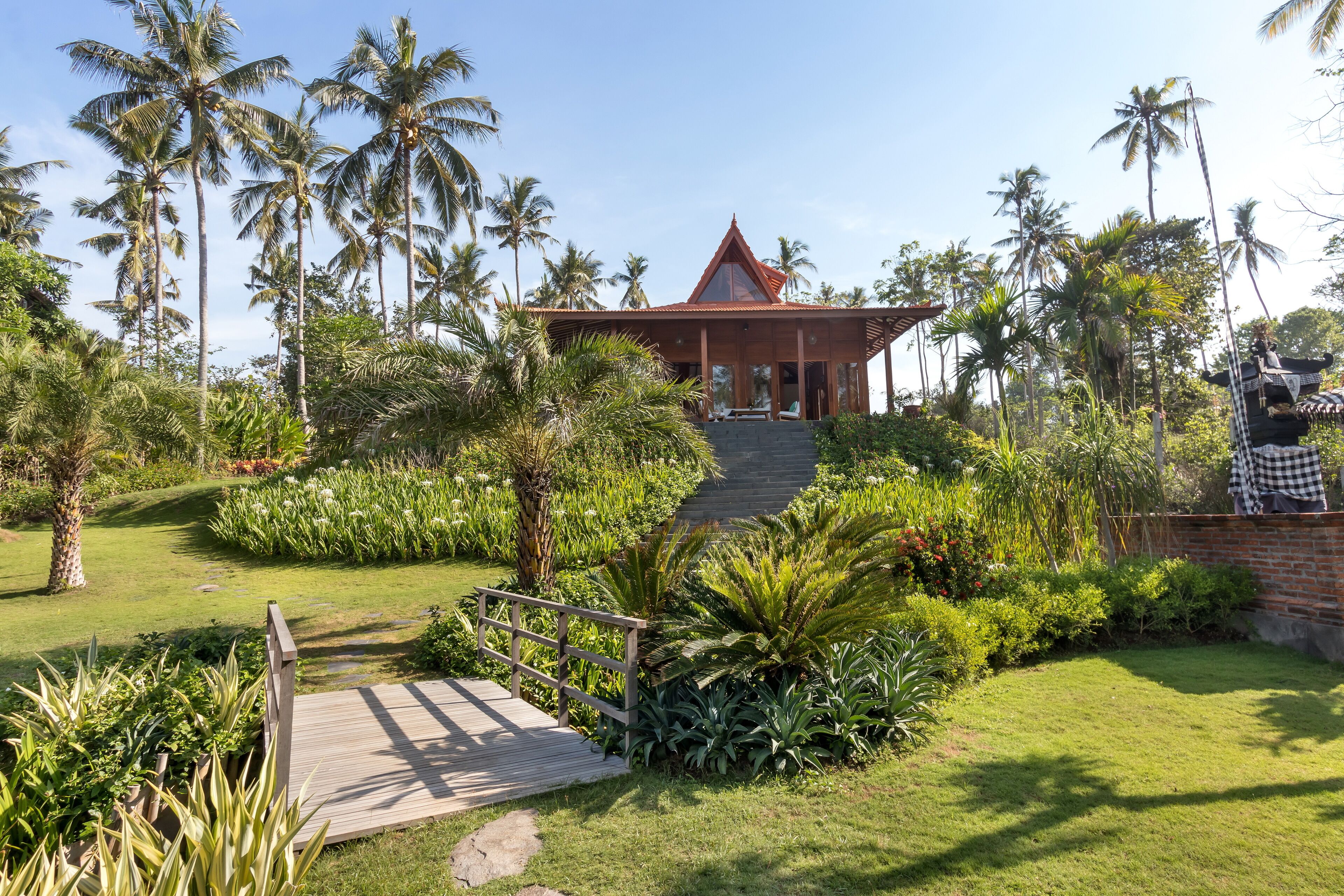 Un resort tropicale con un edificio dal tetto rosso, vegetazione rigogliosa e un ponte di legno che conduce all'ingresso.