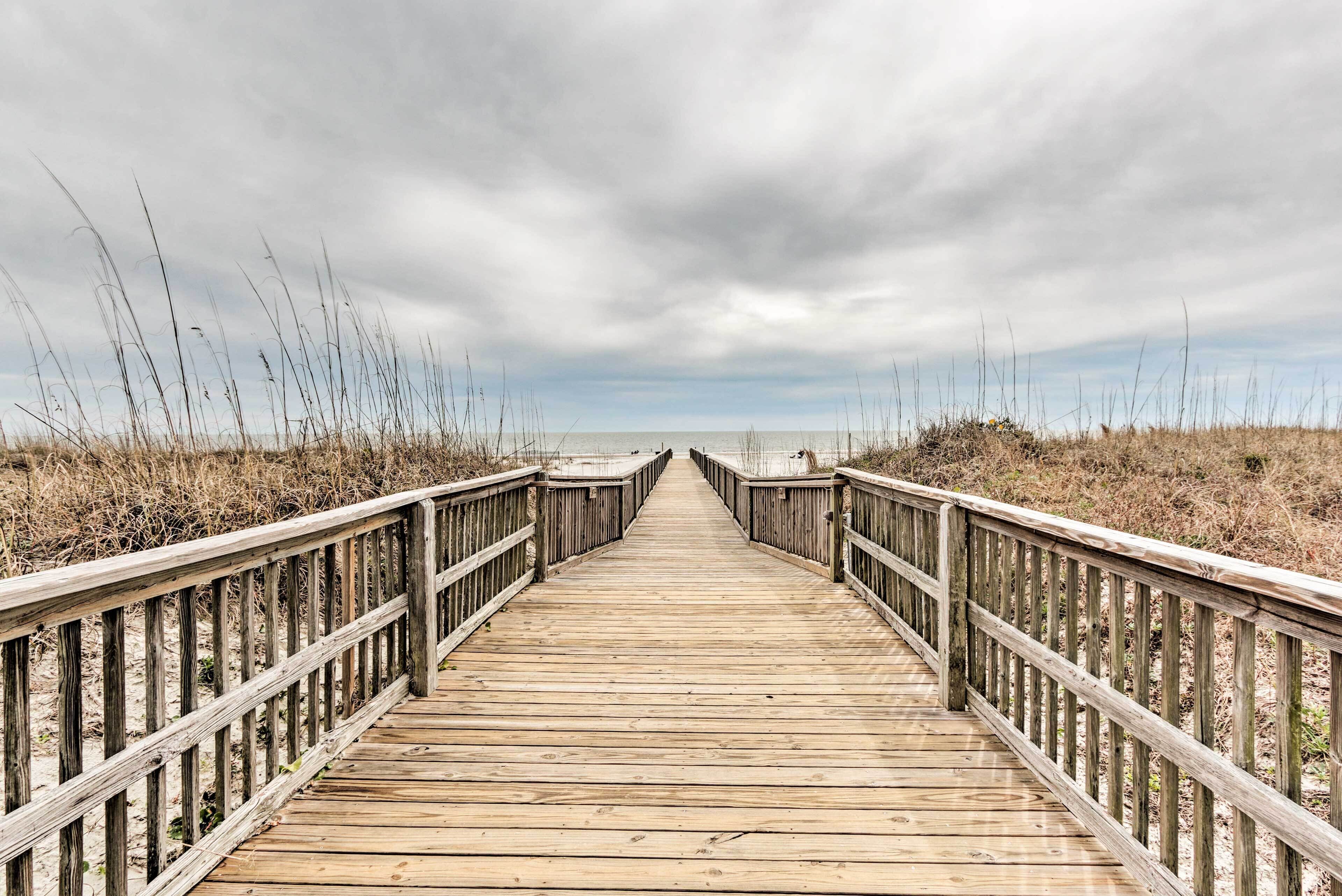 Une promenade en bois menant à la plage, bordée d’herbes hautes et surplombée par un ciel nuageux.