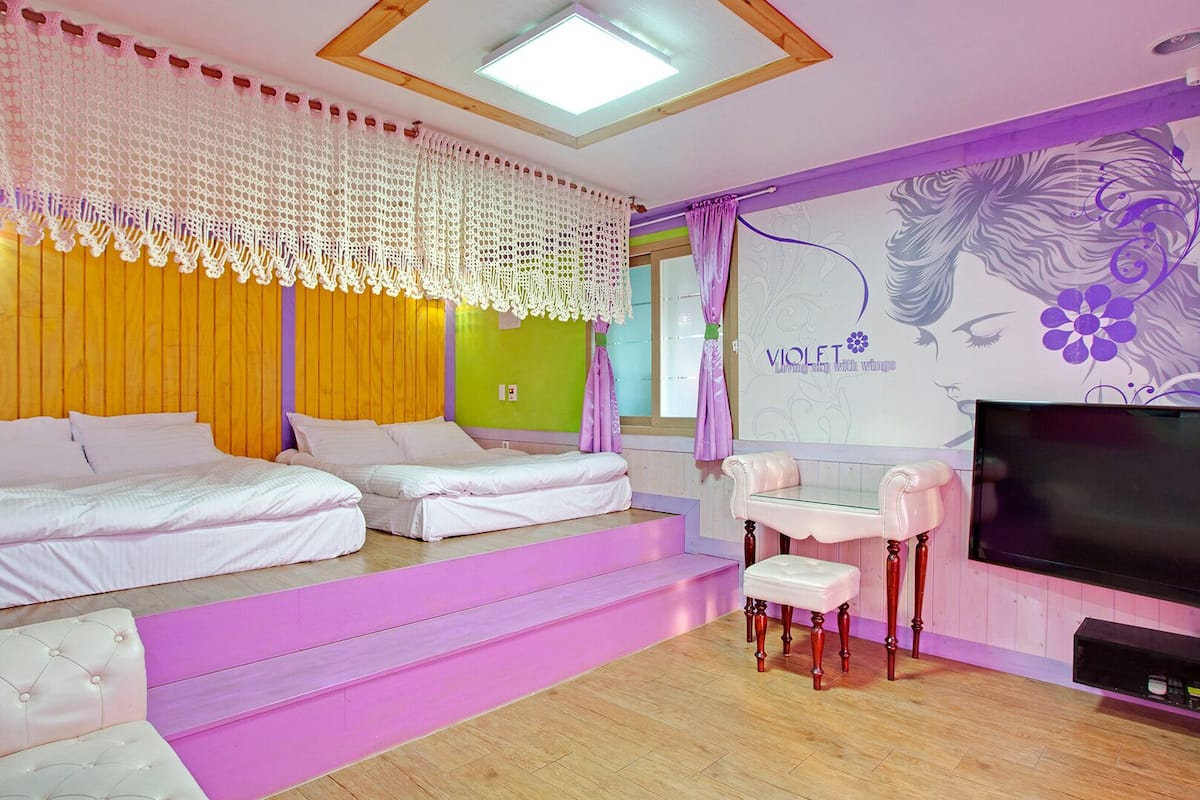 A room with two beds, a vanity with a mirror, a television, and a wall with a floral design.
