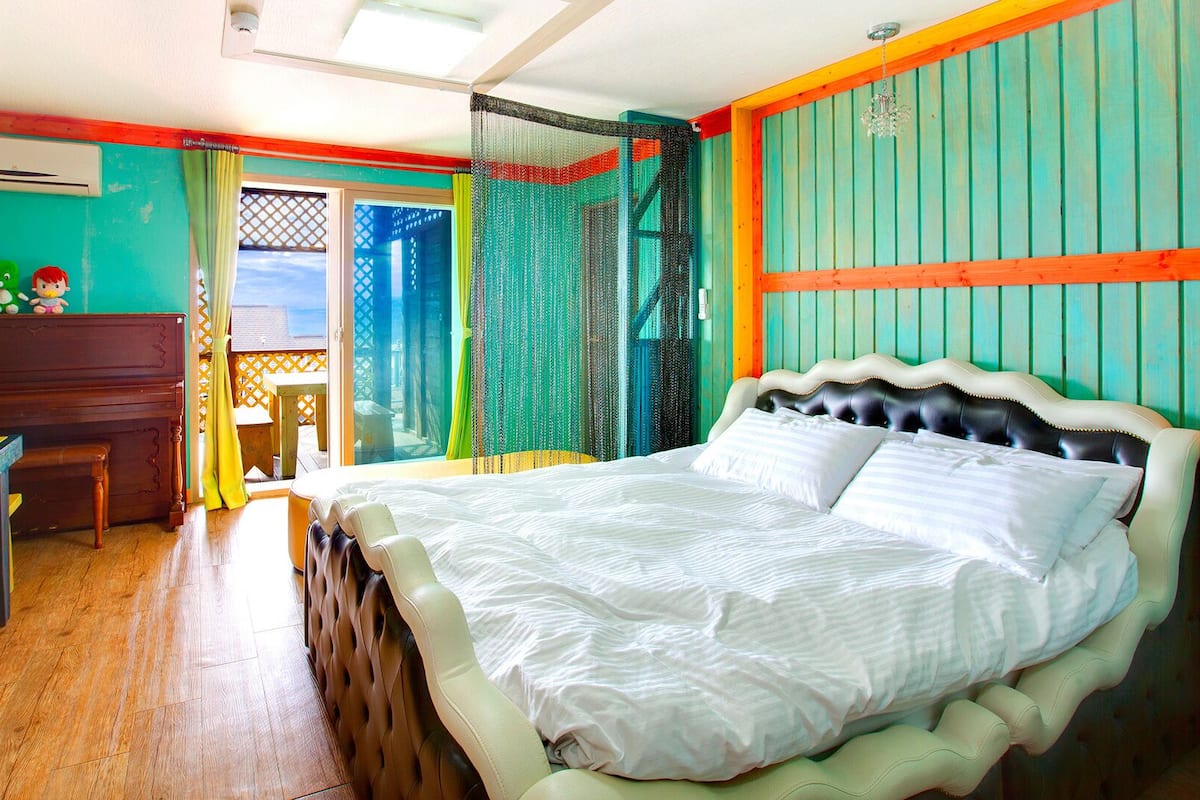 A room with a bed, a desk, and a chair. There is a window with curtains and a view of the outside.