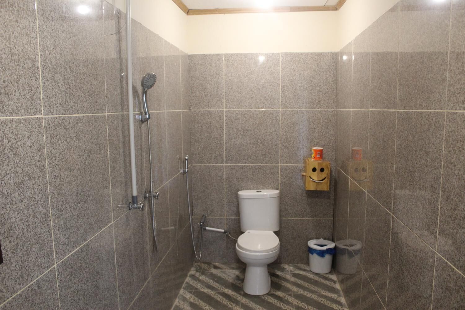 A small, tiled bathroom with a shower, toilet, and a wooden toilet paper holder with a smiley face.