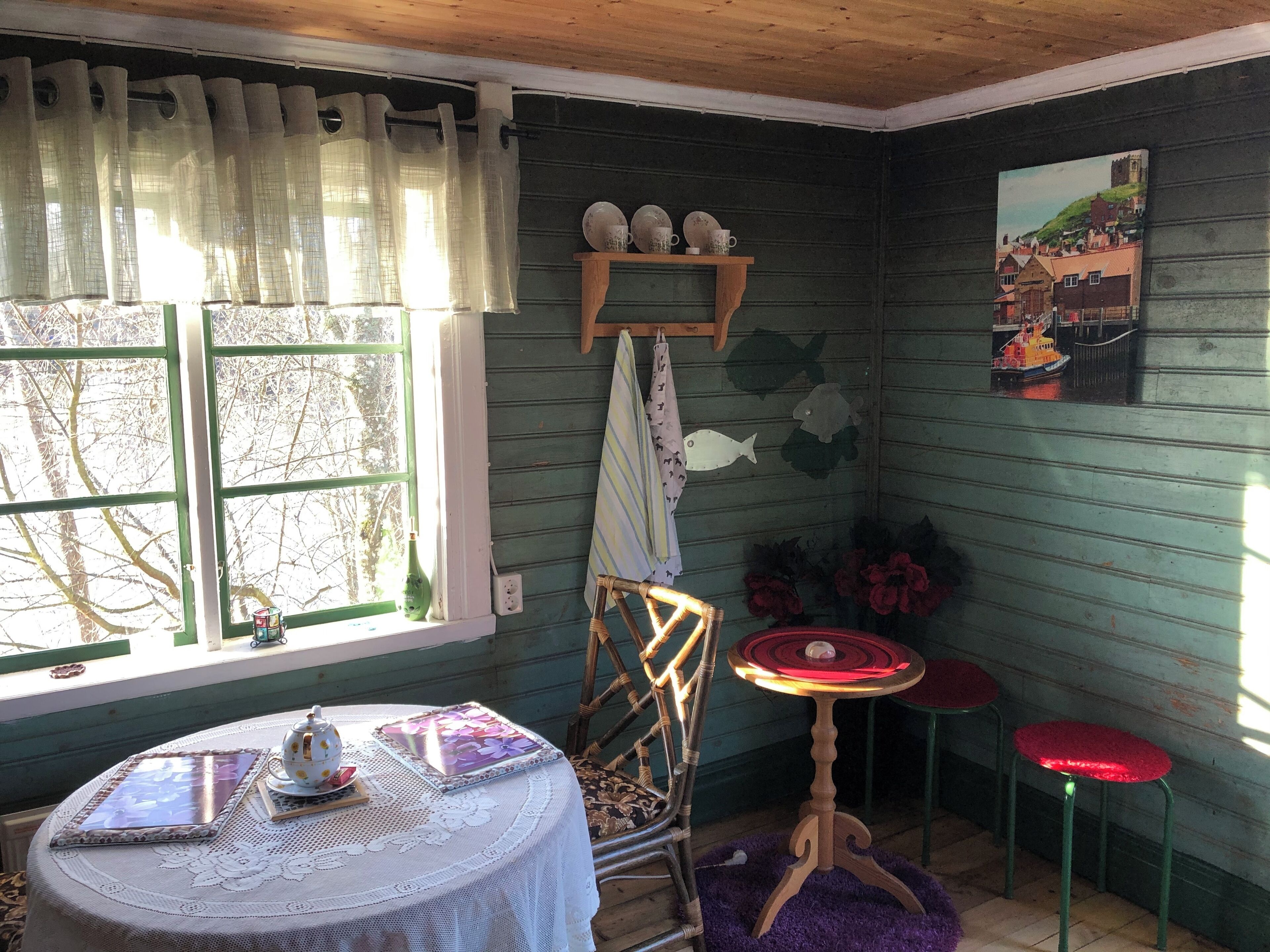 A cozy room with a wooden table, a chair, a small shelf with decorative items, a window with curtains, and a painting on the wall.