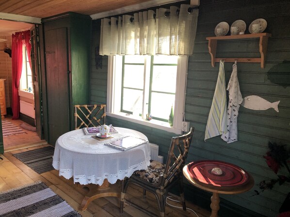 A cozy room with a wooden floor, a round table with a lace tablecloth, a wicker chair, a window with curtains, and a shelf with decorative items.