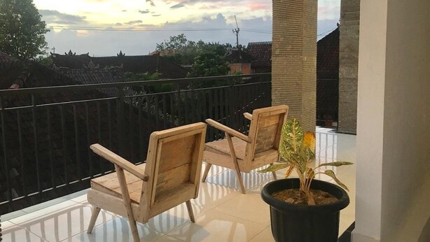 A balcony with two wooden chairs and a potted plant, overlooking a residential area with houses and a clear sky. - Gangga Guesthouse (Canggu)