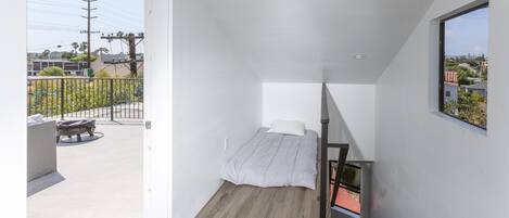 A small, modern bedroom with a single bed, a wooden floor, and a balcony with outdoor furniture.