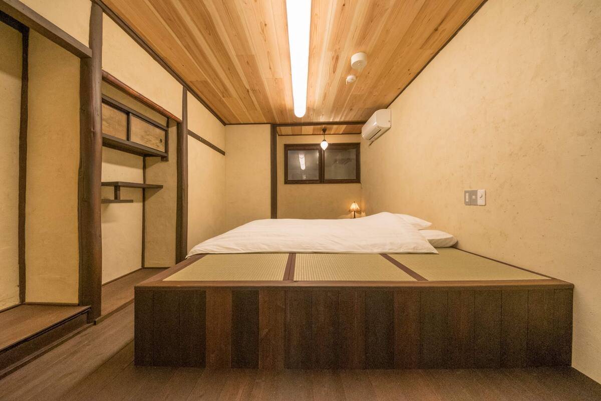 A traditional Japanese-style room with tatami flooring, a low wooden bed, a small window, and a ceiling light fixture.