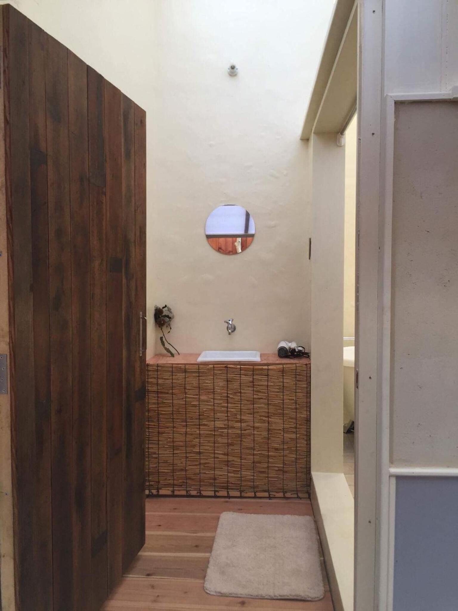 A narrow hallway with a wooden door, a small round mirror, a sink with a towel, and a small rug.