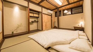 Ett traditionellt japanskt sovrum med tatamimattor på golvet, en stor säng, träbjälkar i taket och ett litet nattduksbord med en lampa.