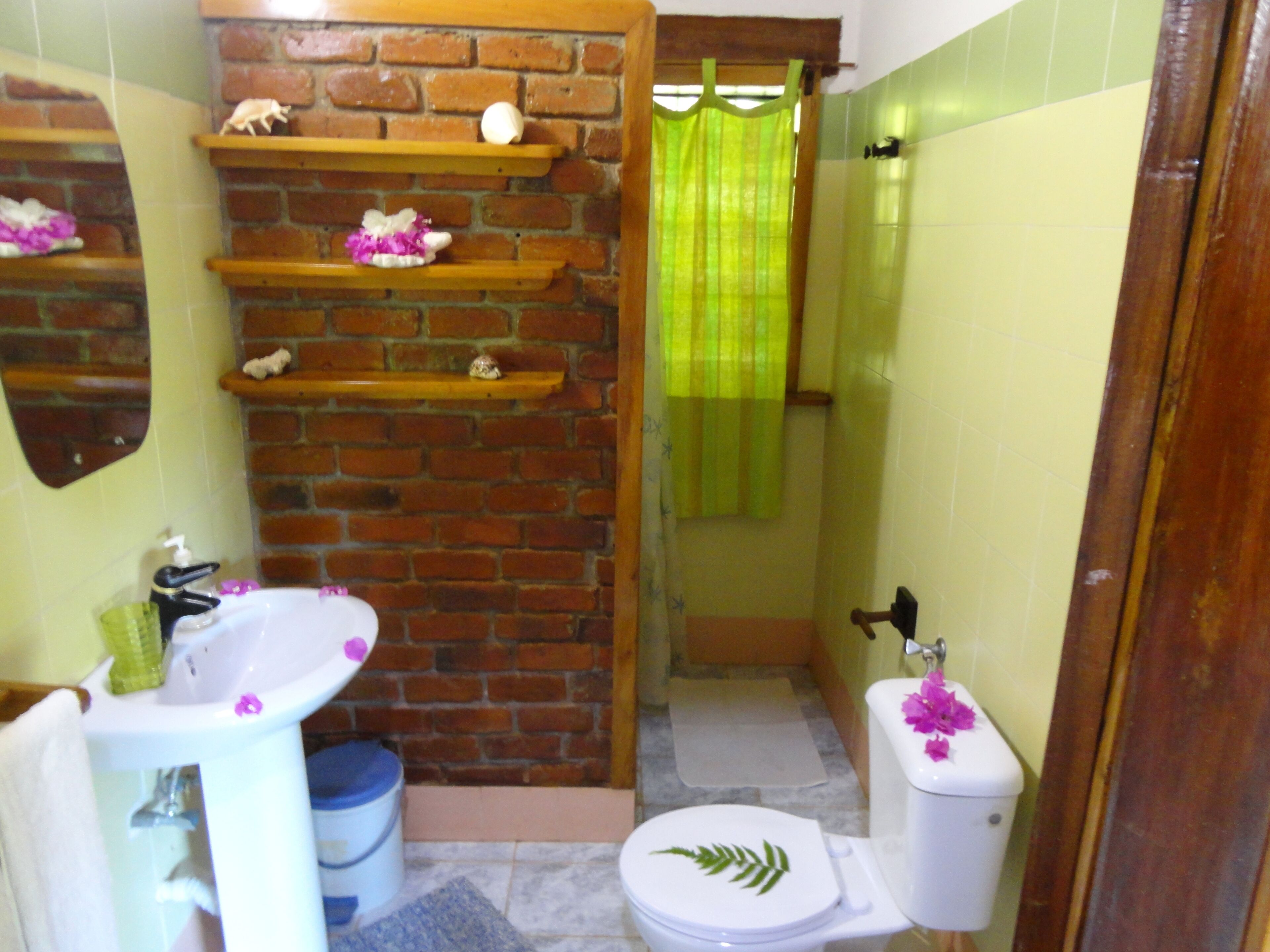 A bathroom with a brick wall, a white sink, a toilet with a leaf decoration, and a shower area with a green curtain.
