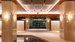 A spacious lobby with a polished floor, wooden columns, and a large glass door leading to a green outdoor area.