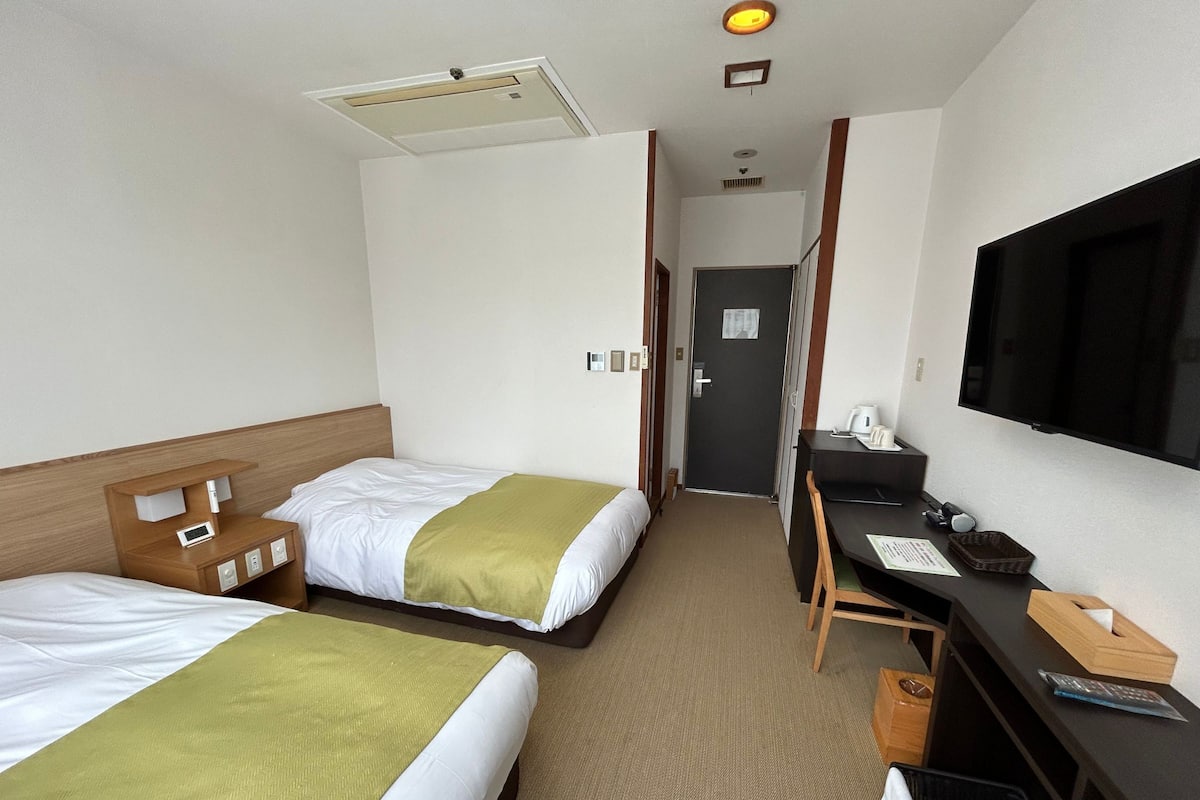 Une chambre d’hôtel avec deux lits, un bureau, une télévision et une porte donnant sur un couloir.