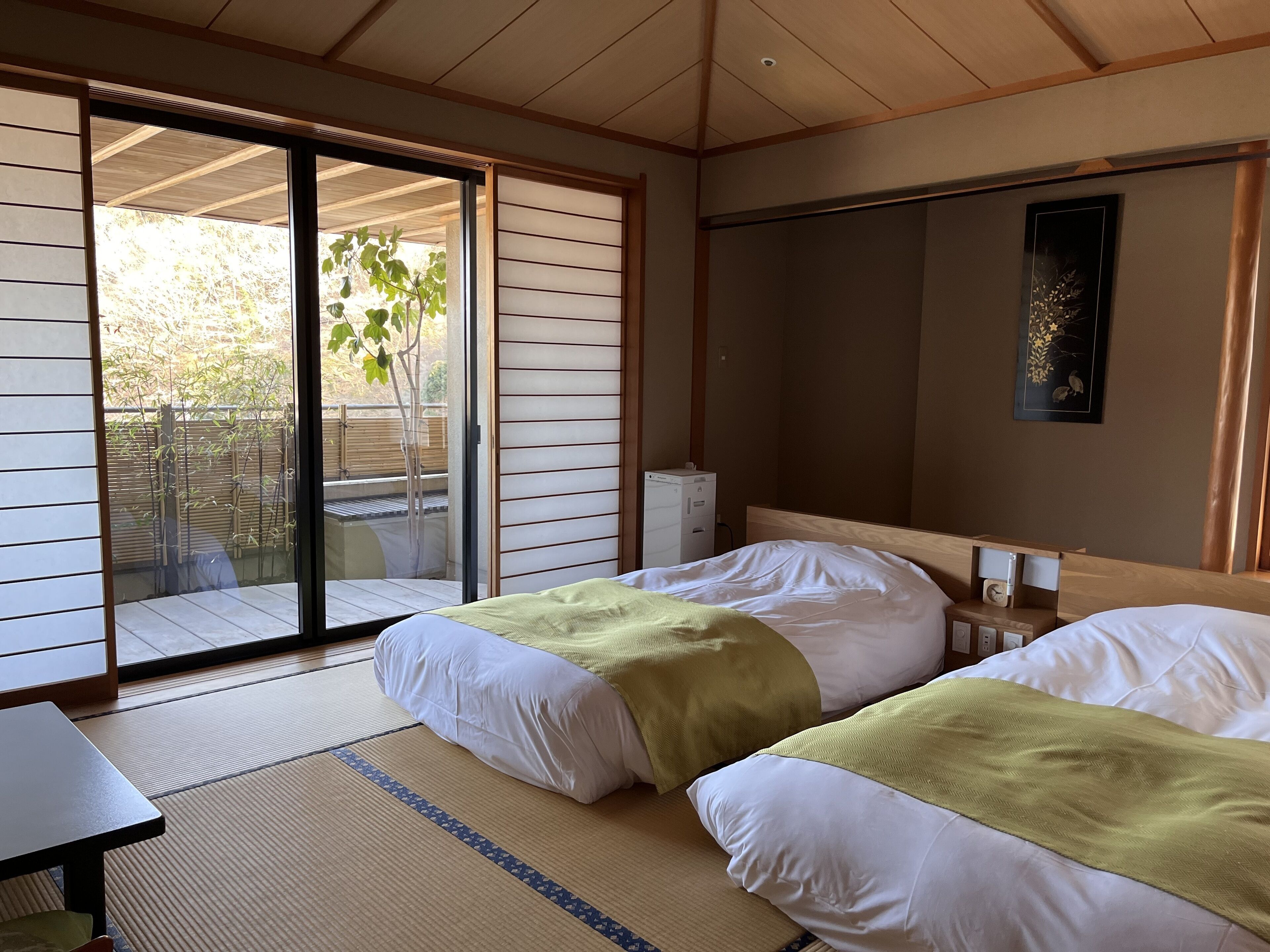 A hotel room with two beds, a sliding glass door to a balcony, a small table, and a wall-mounted clock.