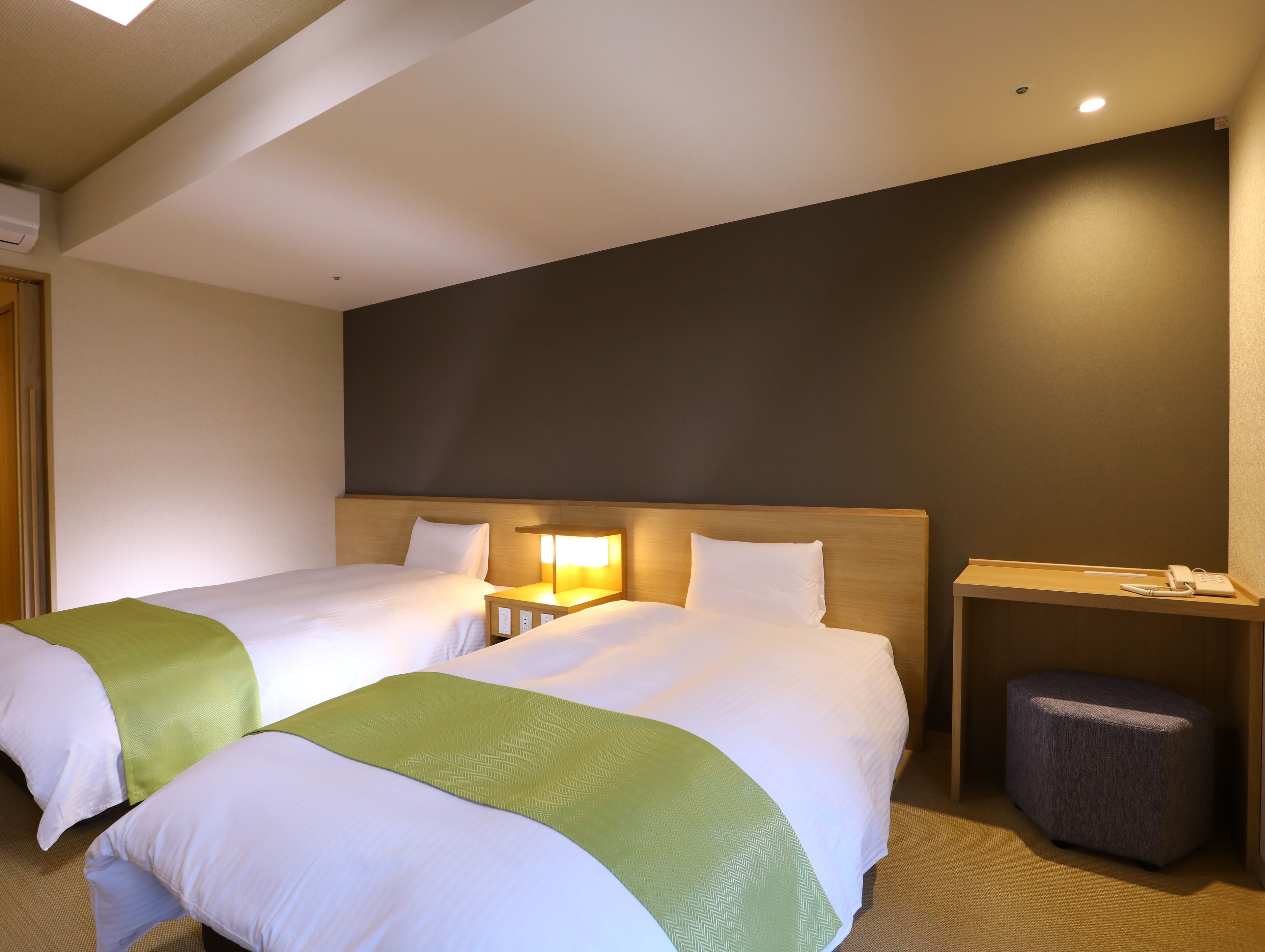 Une chambre d’hôtel avec deux lits, une table de chevet avec une lampe, un petit pouf et un téléphone fixé au mur.