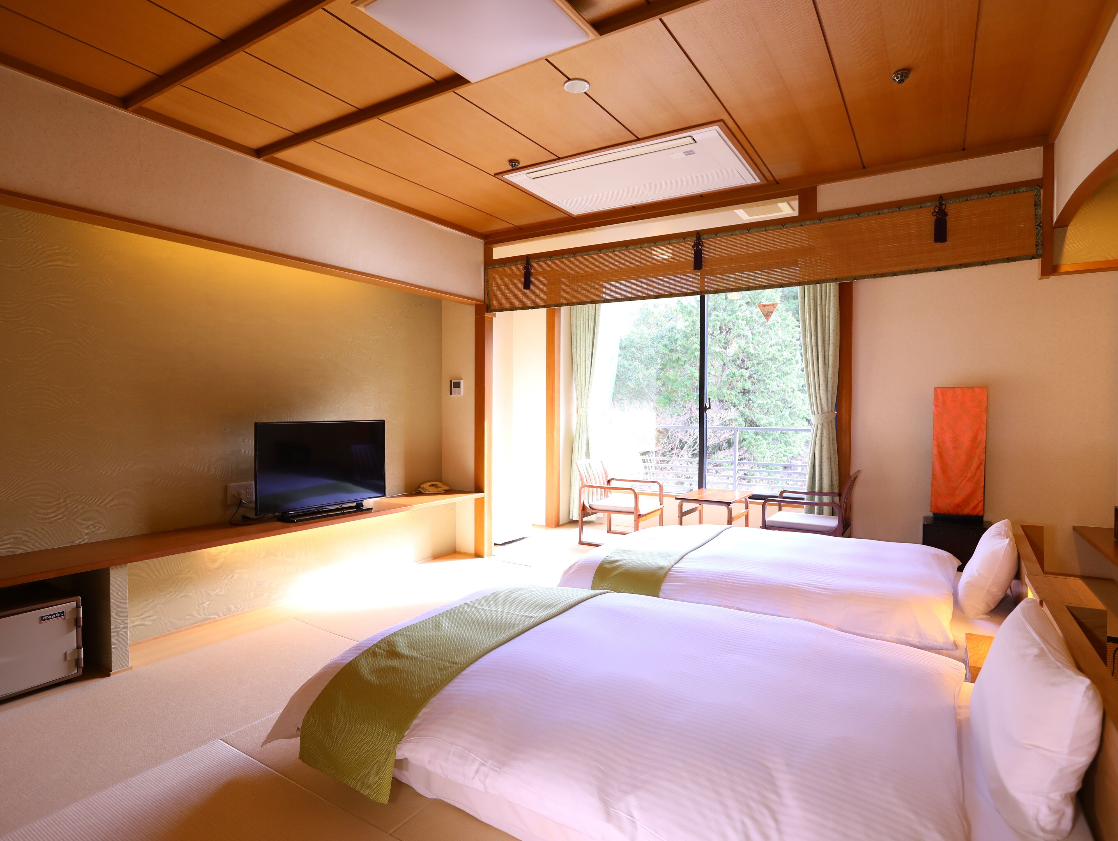 A hotel room with two beds, a television, a desk, and a large window with a view of trees.