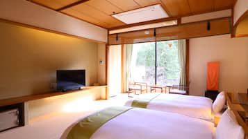 A hotel room with two beds, a television, a desk, and a large window with a view of trees.