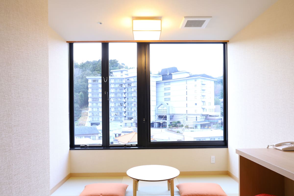 Une pièce avec un sol recouvert de tatamis, une table basse, deux coussins et une grande fenêtre donnant sur des immeubles.
