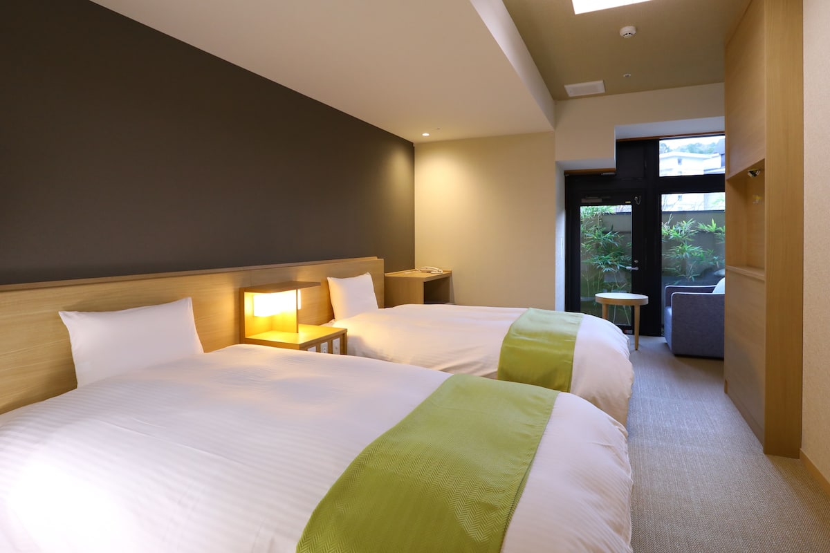 Une chambre d’hôtel avec deux lits, une table de chevet avec une lampe, une petite table et une vue sur l’extérieur.