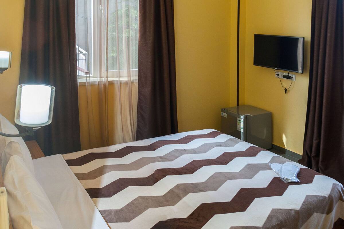 A hotel room with a bed, a television mounted on the wall, a small refrigerator, and a window with curtains.