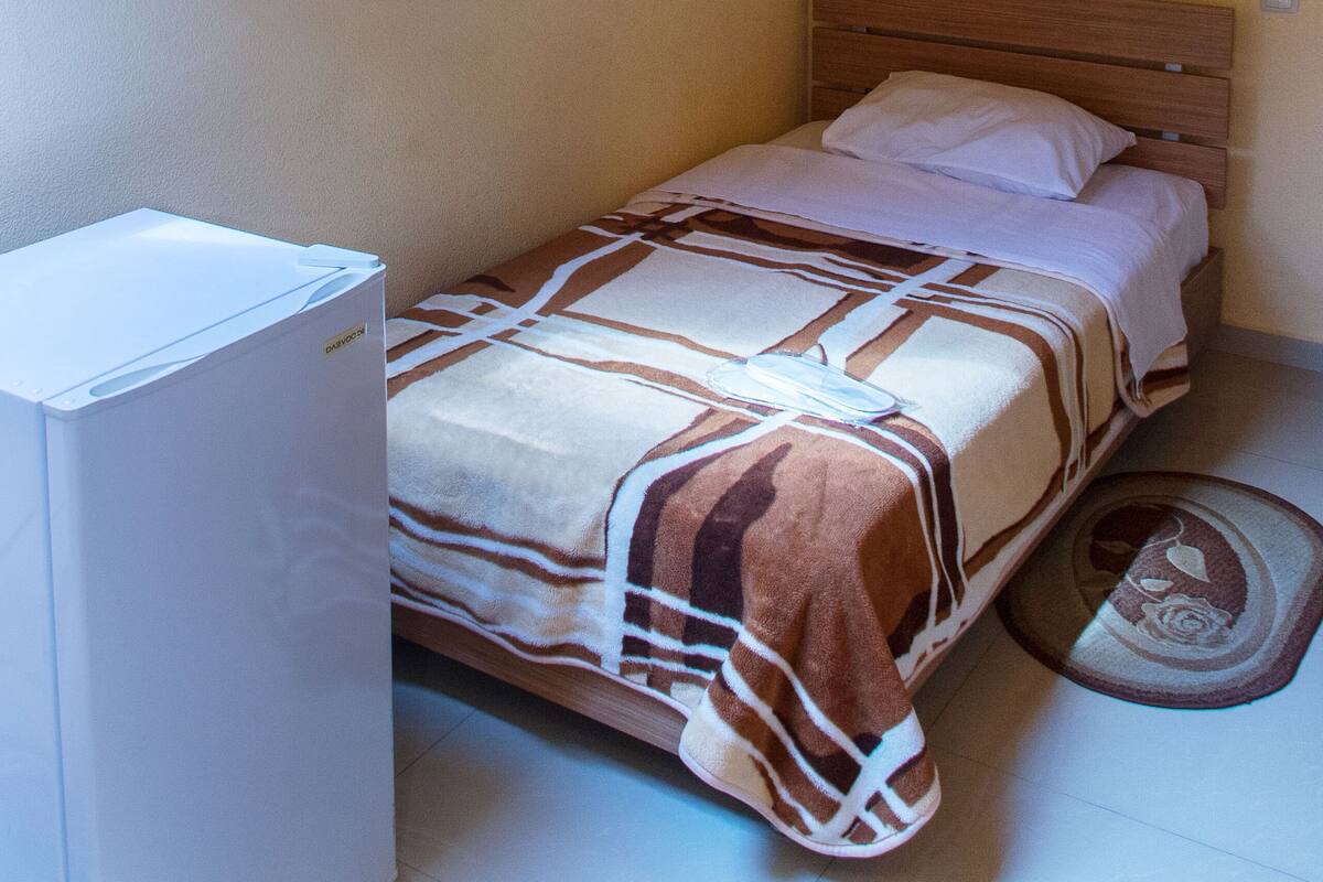 A small, single-bed room with a wooden ceiling, a bed with a patterned blanket, a bedside table, and a small rug.