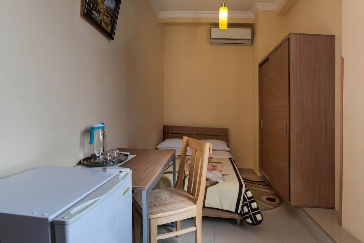 A compact room with a bed, a small table with a kettle, a chair, a wardrobe, and an air conditioning unit.