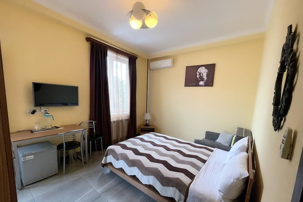 A hotel room with a bed, a desk, a chair, a television, and an air conditioning unit.