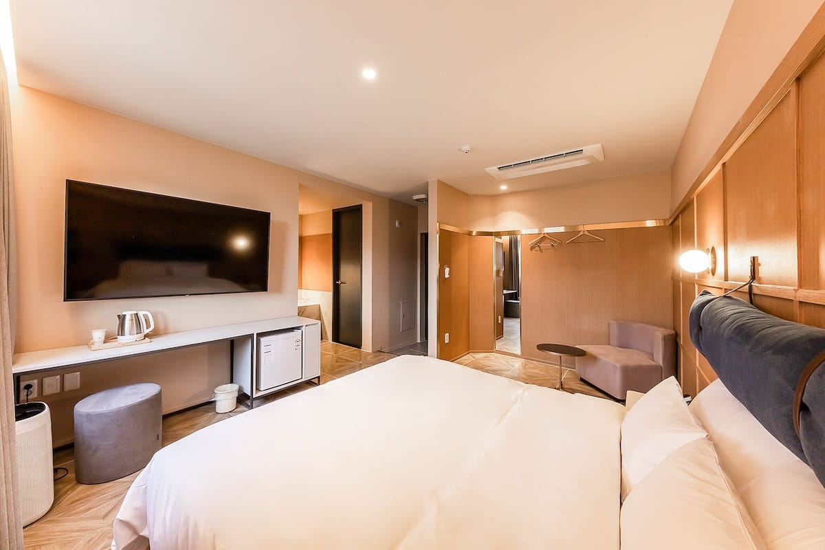 A modern hotel room with a large bed, a flat-screen TV, a desk with a kettle, and a seating area with a chair and table.