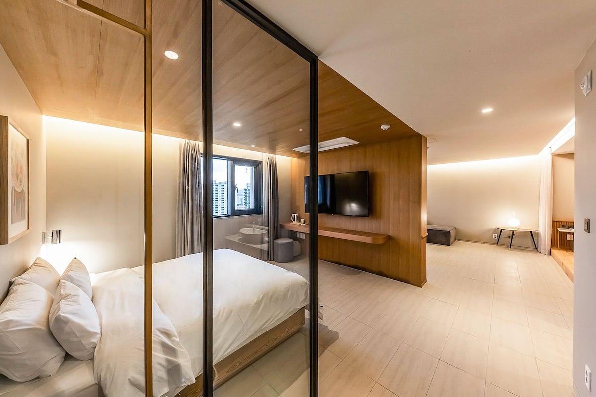 A modern hotel room with a bed, a TV mounted on the wall, a small table, and a bathroom with a sink and mirror.