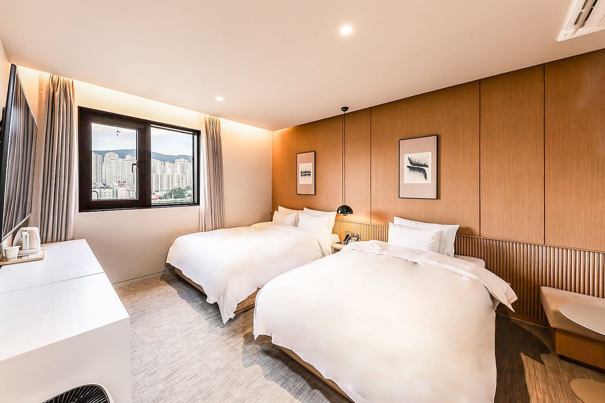 A hotel room with two beds, a desk, a chair, a small table, and a window with a city view.