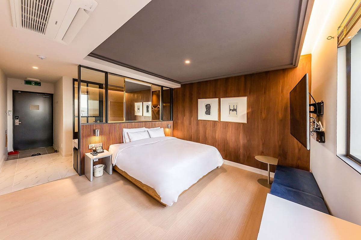 A modern hotel room with a large bed, a bedside table, a small stool, a mirror, and framed artwork on the wall.