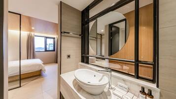 A modern bathroom with a large mirror, a white sink, and a bed in the adjacent room.