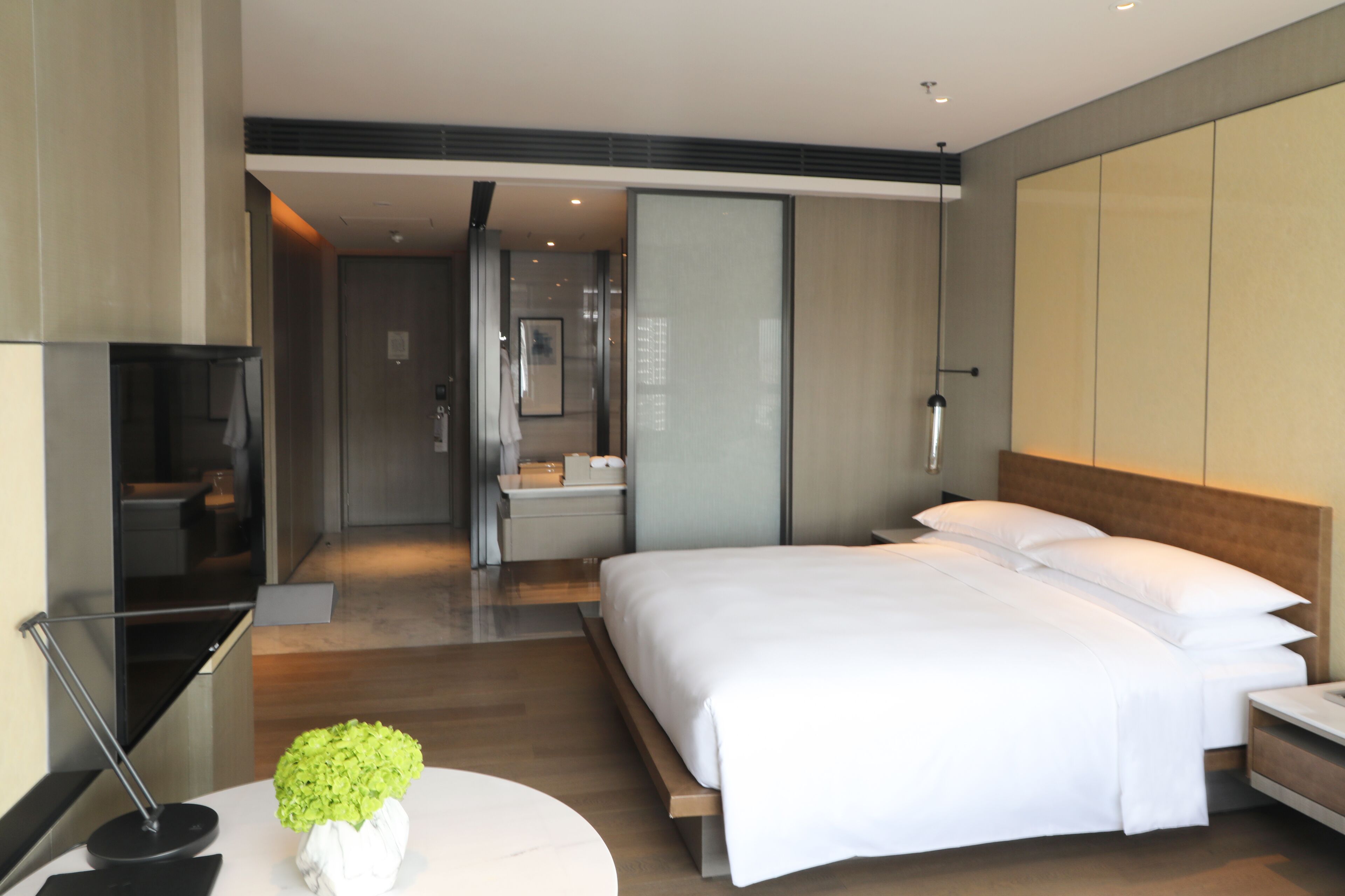 A modern hotel room with a large bed, a bedside table, a TV, and a bathroom visible through an open door.