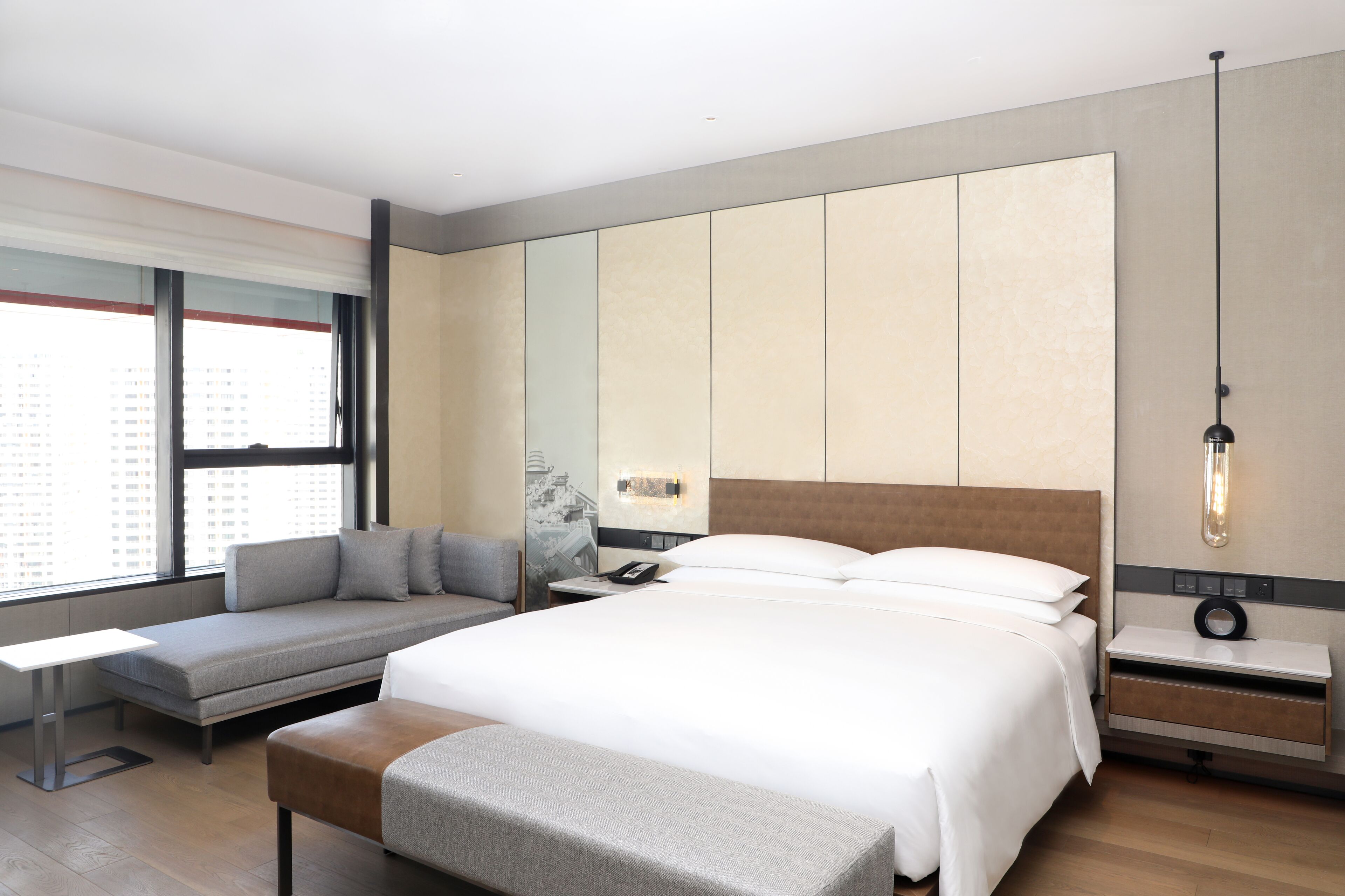 A modern hotel room with a large bed, a seating area, a bedside table, and a large window.