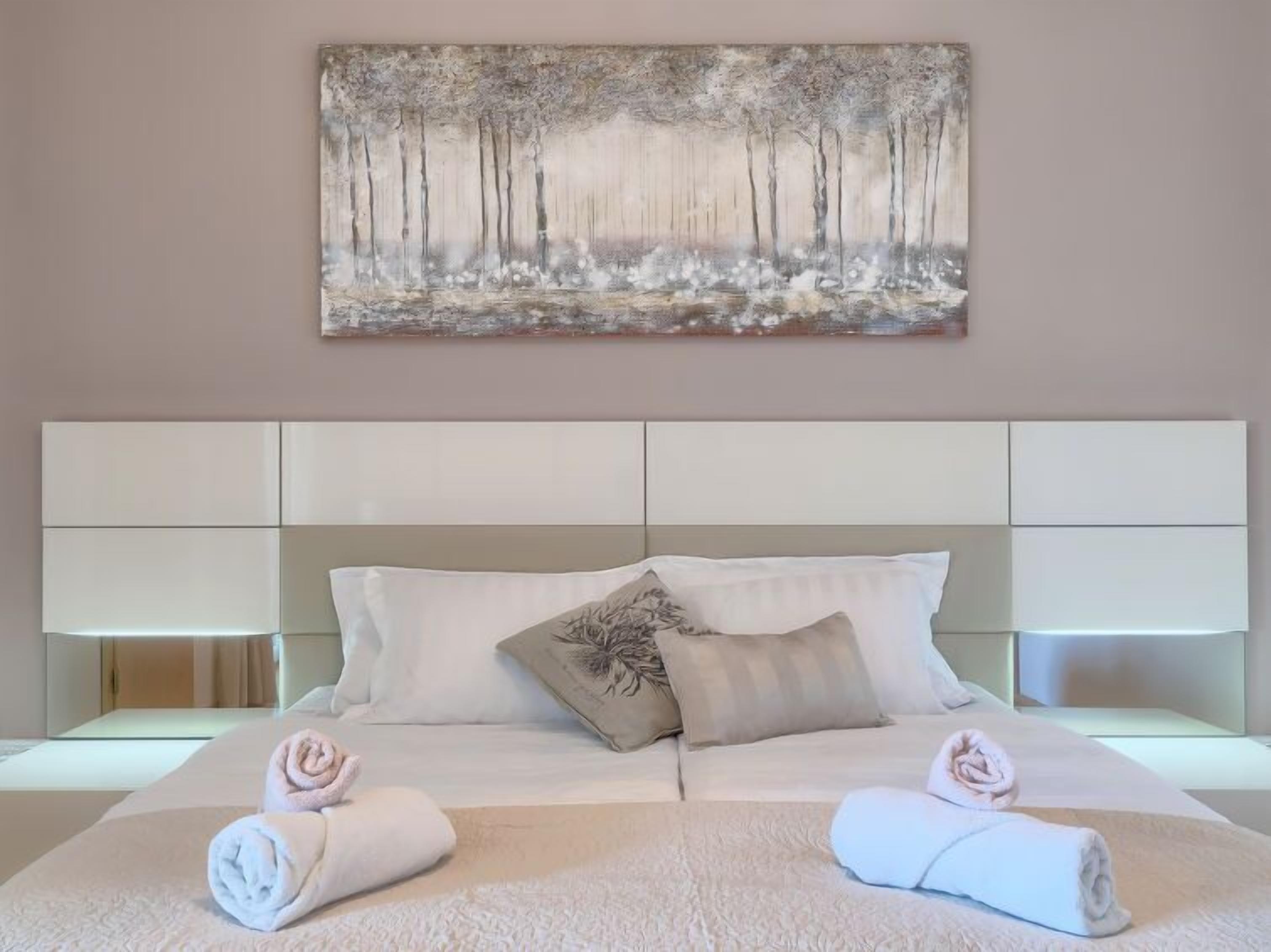 A modern hotel room with a large bed, white linens, two bedside lamps, and a forest-themed painting on the wall.