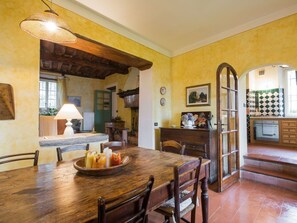 A rustic dining area with a wooden table, chairs, and a bowl of food. There is a lamp, a window, and a view of the kitchen in the background.