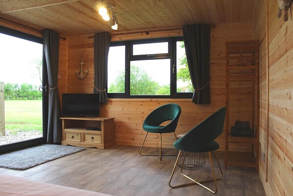 A wooden cabin room with a TV, two chairs, a small table, and a window with curtains.
