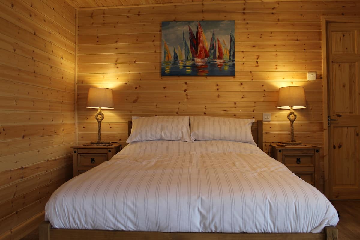 A wooden-paneled room with a bed, two bedside tables with lamps, and a painting of sailboats on the wall.