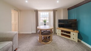 A room with a round wooden table, a television on a wooden stand, a beige sofa, and a window with curtains. - Library View Apartment -  Grade II Listed town centre apartment with parking (Kendal)
