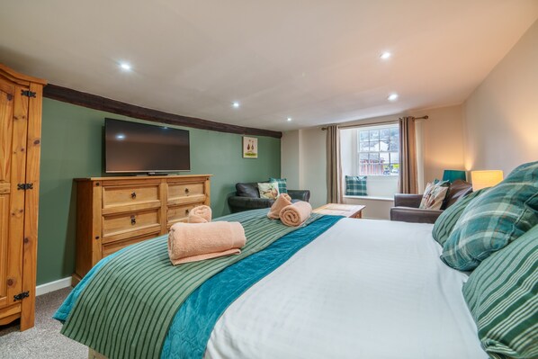 A hotel room with a large bed, a wooden wardrobe, a flat-screen TV, a window with curtains, and a small table with a lamp.