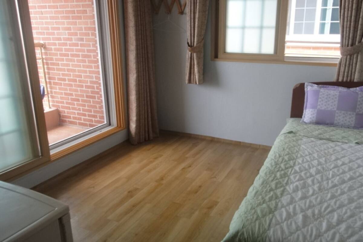 Quarto com piso de madeira, cama com colcha, duas janelas com cortinas e parede de tijolos visível por uma das janelas.